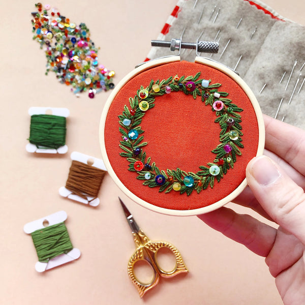 A hand holds an embroidery hoop featuring a green leaf wreath with colorful decorative beads on a red fabric background. The scene includes spools of thread, gold scissors, a pin cushion, and a pile of sequins on a peach-colored surface.