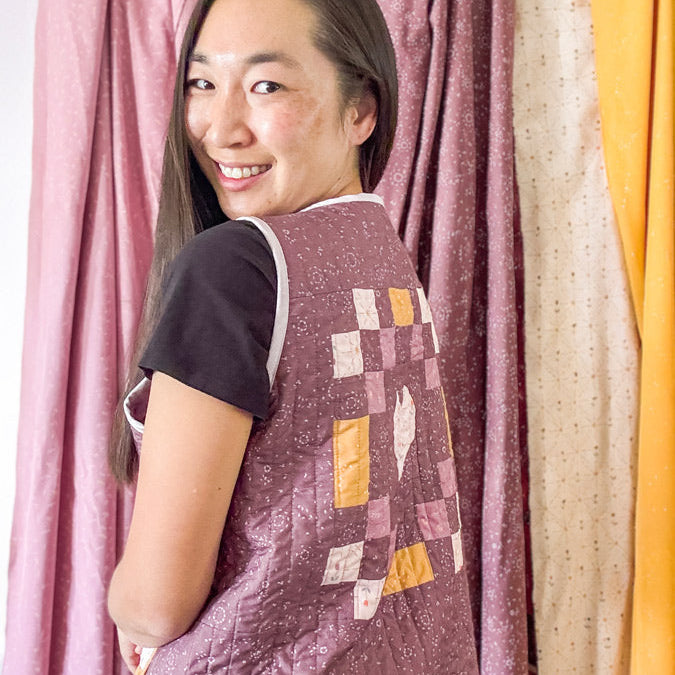 Woman wearing a quilted jacket with a colorful pattern in front of curtains.