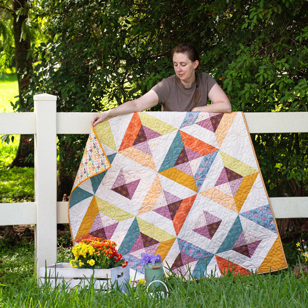 Woman holding a colorful quilt outdoors near a white fence with greenery.