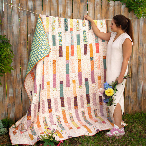 Woman holding a colorful quilt outdoors against a wooden fence.