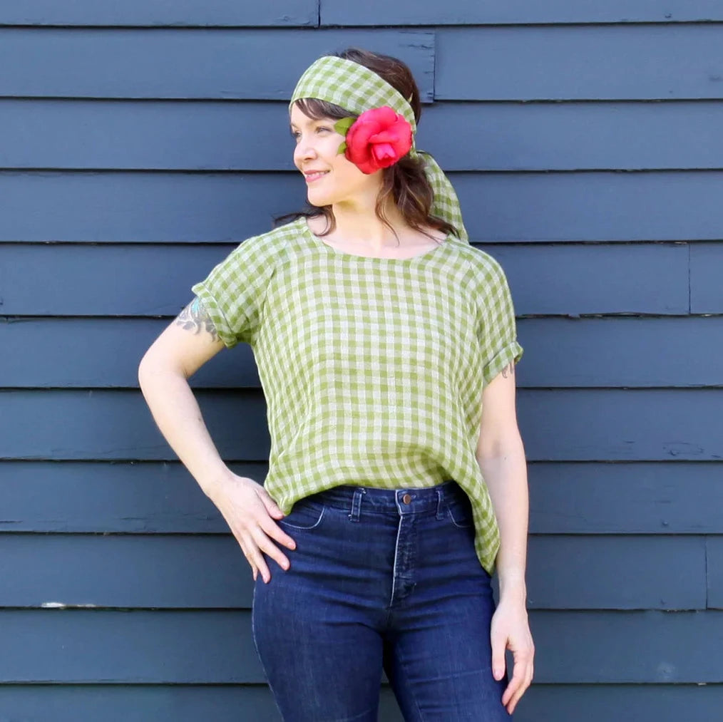 A woman wearing a light green and white gingham short-sleeve top, matching gingham headband, and a large red flower accessory in her hair, posing against a dark blue-gray wooden plank wall.