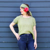 A woman wearing a light green and white gingham short-sleeve top, matching gingham headband, and a large red flower accessory in her hair, posing against a dark blue-gray wooden plank wall.