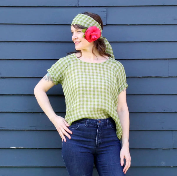 A woman wearing a light green and white gingham short-sleeve top, matching gingham headband, and a large red flower accessory in her hair, posing against a dark blue-gray wooden plank wall.