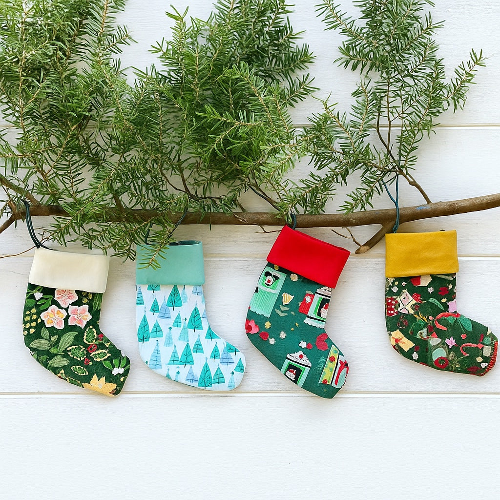 Four colorful Christmas stockings hanging from a branch with greenery on a white wooden surface.