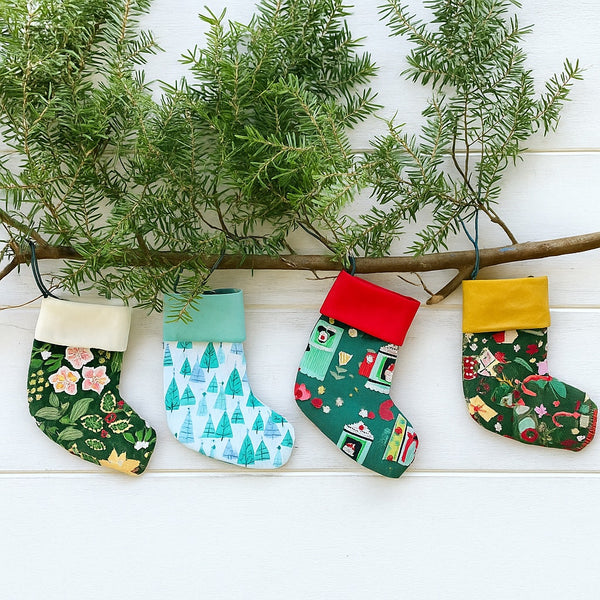 Four colorful Christmas stockings hanging from a branch with greenery on a white wooden surface.